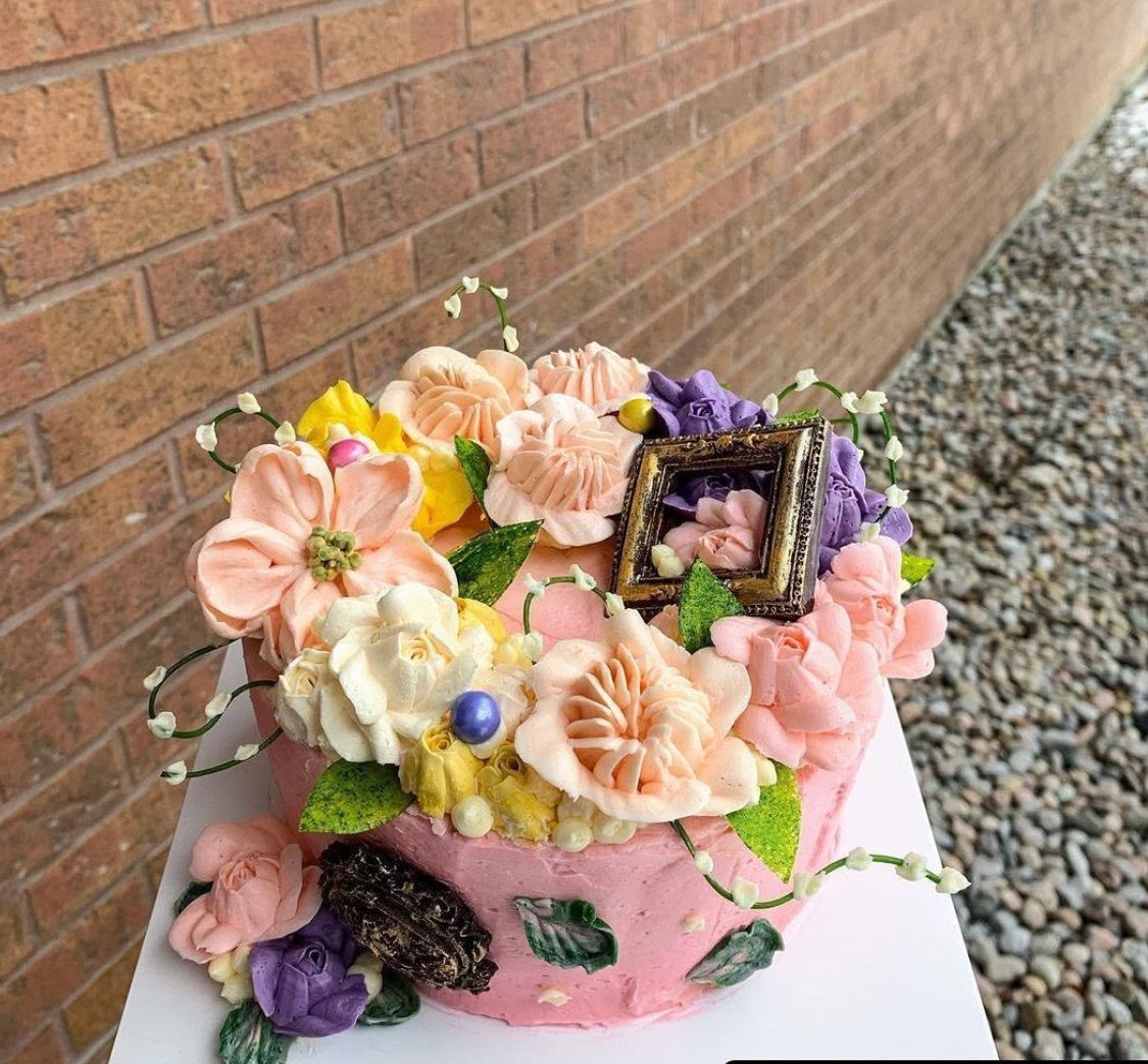 Pink cake with peach, purple and yellow buttercream flowers against a brick wall